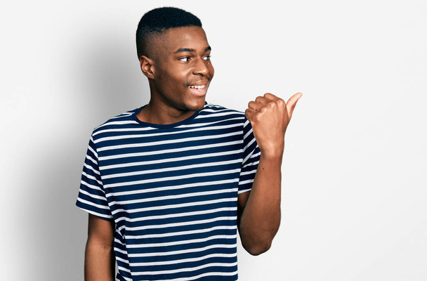 Sıradan çizgili tişörtler giyen genç Afro-Amerikan bir adam başparmağıyla yan tarafı işaret ediyor ve ağzı açık gülümsüyor.  - Fotoğraf, Görsel