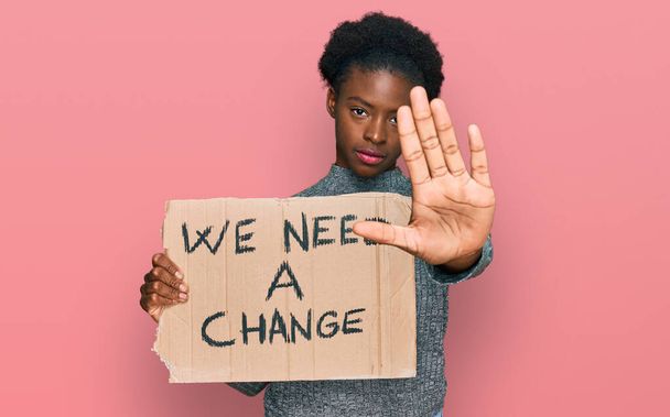 Giovane ragazza afroamericana in possesso abbiamo bisogno di un banner cambiamento con mano aperta facendo stop segno con espressione seria e sicura, gesto di difesa  - Foto, immagini