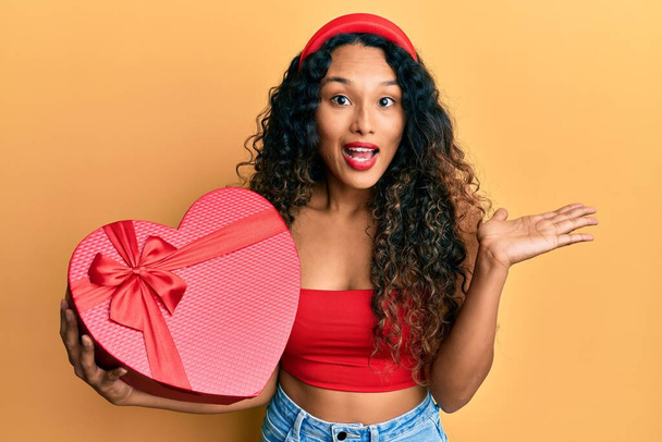 Young latin woman holding valentine day gift celebrating achievement with happy smile and winner expression with raised hand  - Photo, Image