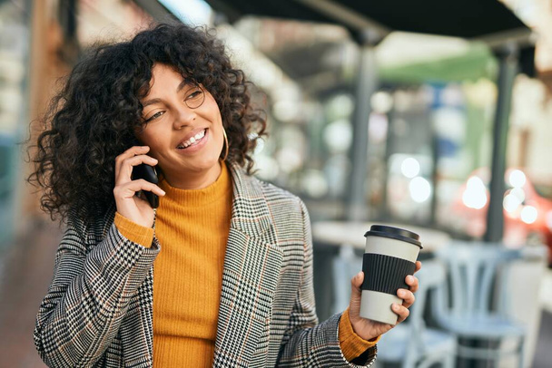 Junge hispanische Geschäftsfrau spricht auf dem Smartphone und trinkt Kaffee in der Stadt. - Foto, Bild