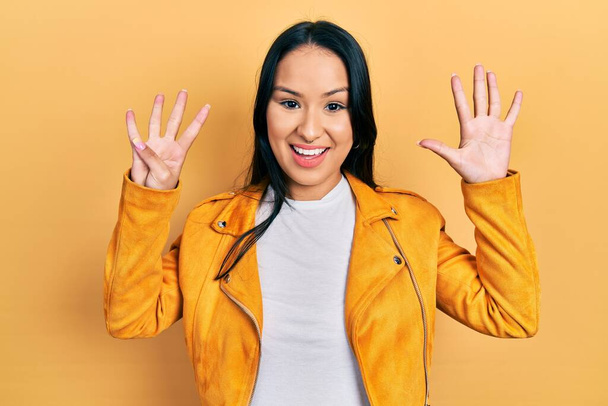 Gyönyörű spanyol nő orrpiercinggel sárga bőrdzsekiben, mutatva és mutatva a kilences számú ujjakkal, miközben magabiztosan és boldogan mosolyog..  - Fotó, kép