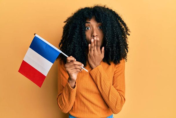 Африканська американка з волоссям афро, що тримає прапор франсу закриваючи рот рукою, шокована і налякана за помилку. здивований вираз  - Фото, зображення