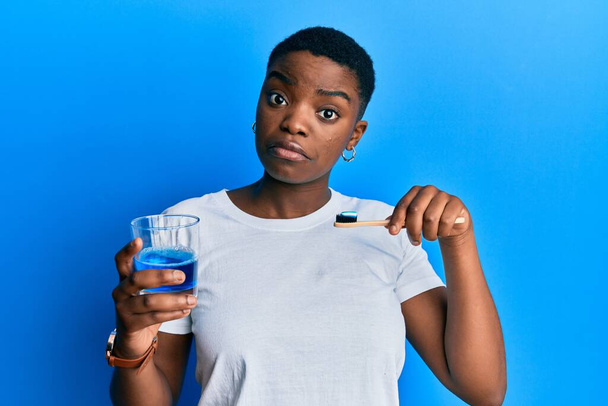 Joven mujer afroamericana sosteniendo un vaso de enjuague bucal y cepillo de dientes para una expresión fresca y confusa. concepto de duda.  - Foto, Imagen
