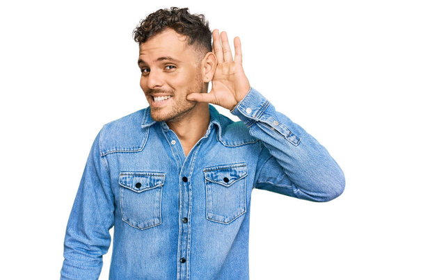 Молодой латиноамериканец в джинсовой куртке, улыбаясь рукой над ухом, слушает слухи и сплетни. концепция глухоты.  - Фото, изображение