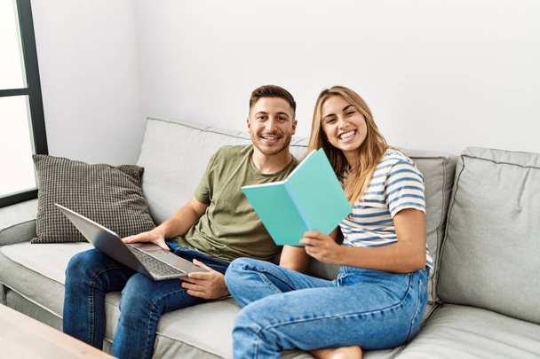 Nuori latino pari käyttää kannettavaa tietokonetta ja lukeminen kirja istuu sohvalla kotona. - Valokuva, kuva