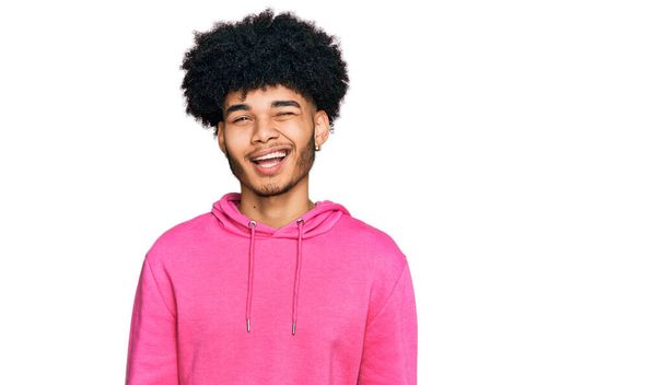 若いですアフリカ系アメリカ人男性とともにafro髪身に着けていますカジュアルピンクのスウェットシャツウインク見ますザカメラとともにセクシー式,陽気で幸せな顔.  - 写真・画像