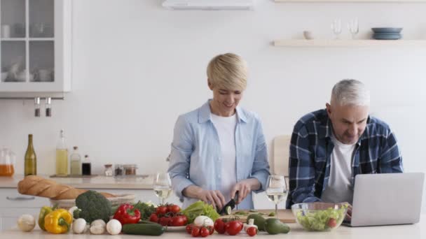 Reif ehemann using laptop während ehefrau preparing dinner im küche - Filmmaterial, Video