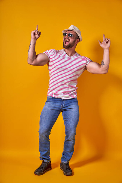 Portrait of optimistic funky guy dance wear summer look straw hat, sunglasses isolated on yellow color background - 写真・画像