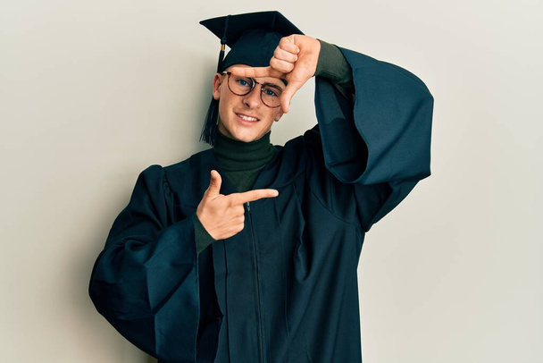 卒業キャップと式のローブを着た若い白人男性が、幸せそうな顔をした手と指でフレームを作って笑っています。創造性と写真の概念.  - 写真・画像