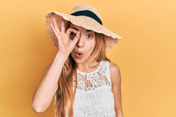 Młoda biała kobieta w letniej czapce robi ok gest wstrząśnięty zaskoczoną twarzą, okiem patrzącym przez palce. niewierząca ekspresja.  - Zdjęcie, obraz
