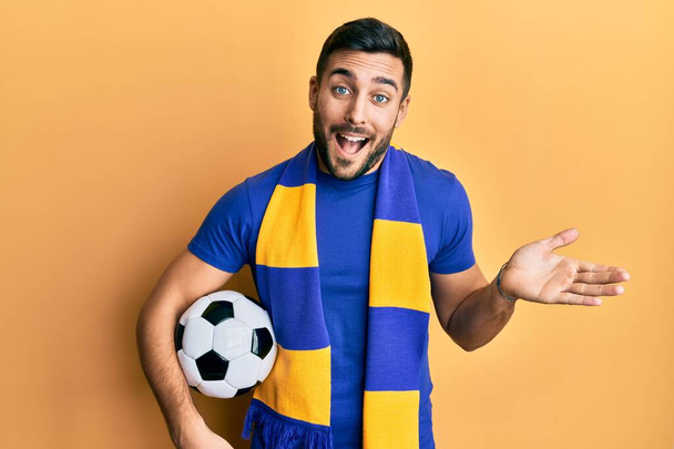 Mladý hispánec fotbalový chuligán fotbalový míč slaví úspěch se šťastným úsměvem a vítězným výrazem se zvednutou rukou  - Fotografie, Obrázek