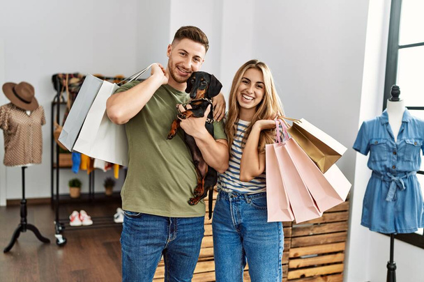 Joven pareja de clientes hispanos sonriendo feliz sosteniendo bolsas de compras y perros en la tienda de ropa. - Foto, Imagen