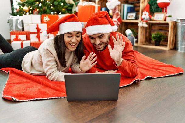 Young hispanic couple having video call using laptop lying on the floor by christmas tree at home. - Photo, Image