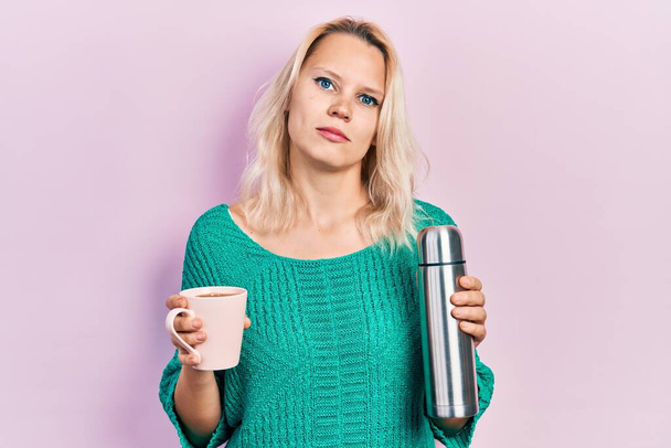 Beautiful caucasian blonde woman drinking a cup of coffee holding thermo relaxed with serious expression on face. simple and natural looking at the camera.  - Photo, Image