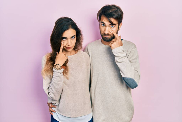 Молодая испаноязычная пара в повседневной одежде, указывая на глаза, наблюдая за твоим жестом, подозрительным выражением лица.  - Фото, изображение
