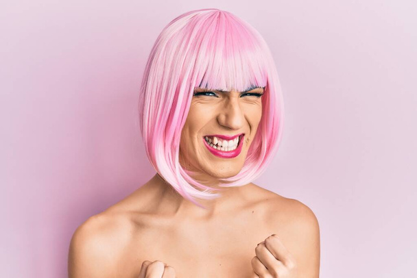 Молодой человек в женском макияже надевает розовый парик, радуясь успеху с поднятыми руками и закрытыми глазами, радуясь победе. победитель концепция.  - Фото, изображение