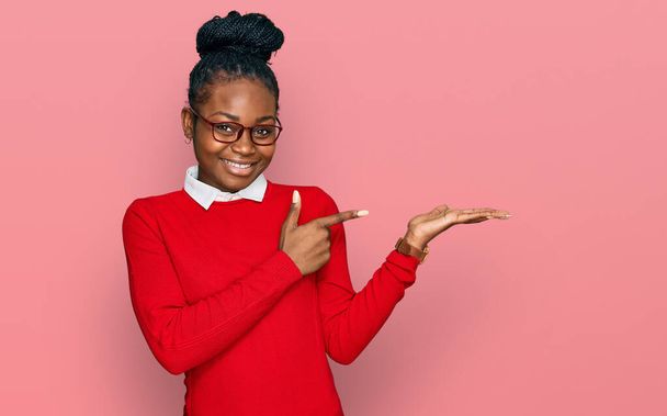 Mladá afroameričanka v neformálním oblečení a brýlích užaslá a usmívající se na fotoaparát při prezentaci rukou a ukazování prstem.  - Fotografie, Obrázek