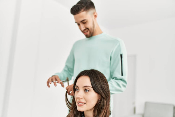 Young hispanic stylist man cutting woman's hair using scissors at beauty center. - Foto, immagini