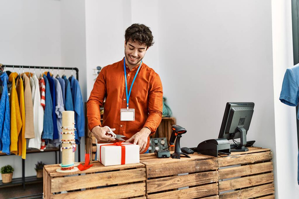 młody Latynos sklepikarz człowiek uśmiechnięty szczęśliwy przygotowując prezent w sklepie odzieżowym. - Zdjęcie, obraz