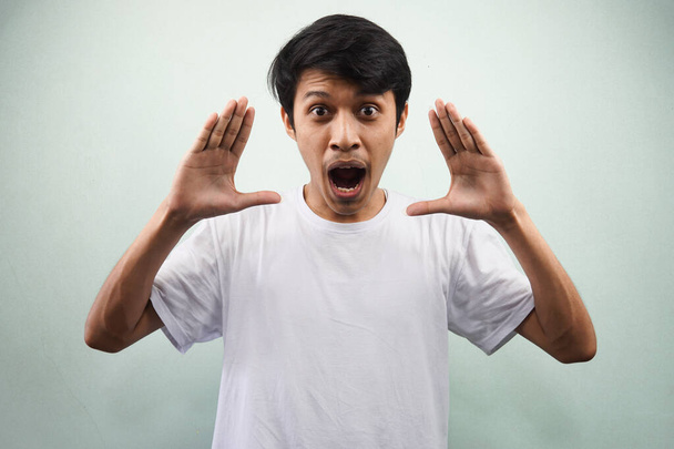 Молодий азіатський чоловік у білій футболці кричить перед камерою з обома руками, піднятими ізольовано на сірому фоні
 - Фото, зображення