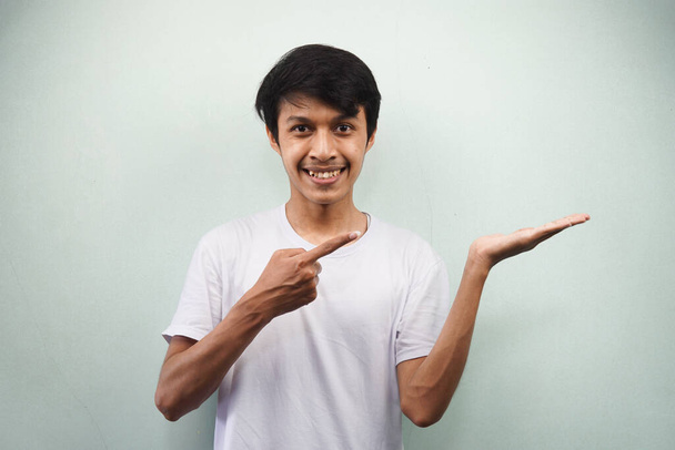 Junger asiatischer Mann in weißem T-Shirt mit lächelndem und selbstbewusstem Gesichtsausdruck, der in die Kamera blickt und mit der Hand auf den leeren Raum zeigt, der ein auf grauem Hintergrund isoliertes Produkt zeigt, Werbekonzept - Foto, Bild
