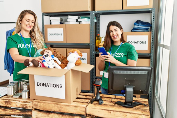 Duas jovens voluntárias sorrindo felizes trabalhando no centro de caridade. Menina sorrindo feliz usando smartphone. - Foto, Imagem