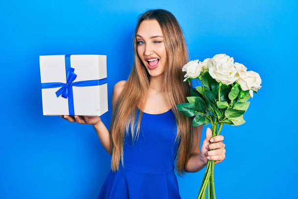 Junges blondes Mädchen mit Geburtstagsgeschenk und Blumenstrauß zwinkert in die Kamera mit sexy Ausdruck, fröhlichem und glücklichem Gesicht.  - Foto, Bild