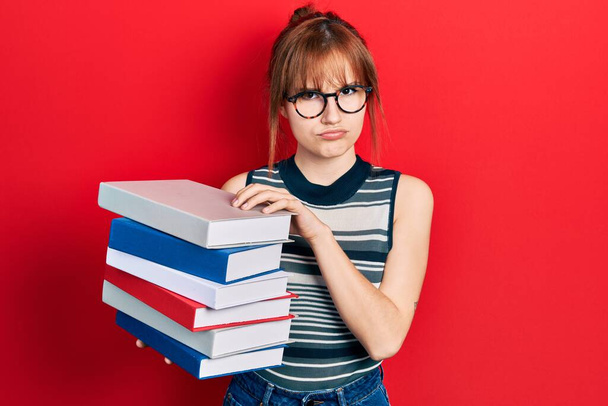 Jeune femme rousse tenant une pile de livres sceptiques et nerveux, fronçant les sourcils bouleversés à cause de problèmes. personne négative.  - Photo, image