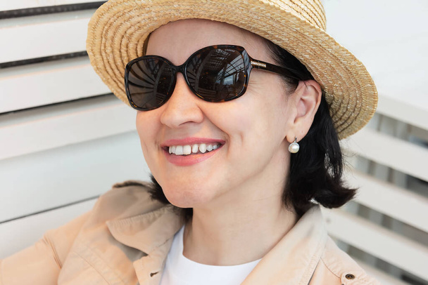 Портрет улыбающейся молодой женщины в летней одежде, солнцезащитных очках и соломенной шляпе, сидящей на летней террасе кафе - Фото, изображение