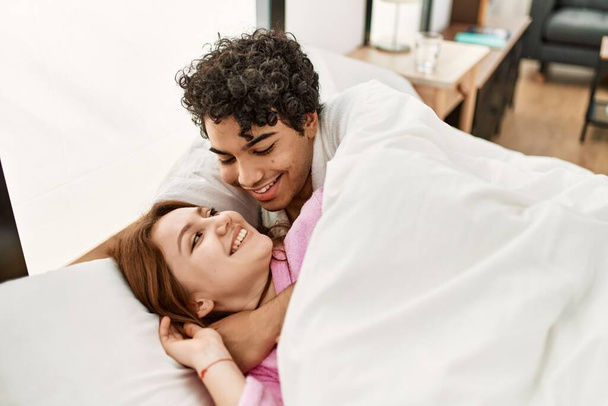 Νεαρό ζευγάρι χαμογελά ευτυχισμένη και αγκαλιάζει ξαπλωμένος στο κρεβάτι στο υπνοδωμάτιο. - Φωτογραφία, εικόνα