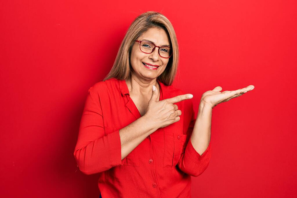 Hispanische Frau mittleren Alters in lässiger Kleidung und Brille staunt und lächelt in die Kamera, während sie mit der Hand präsentiert und mit dem Finger zeigt.  - Foto, Bild
