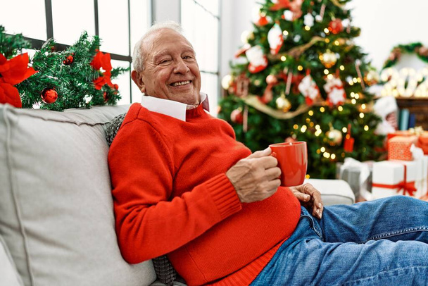 Idősebb ősz hajú férfi kávét iszik, ünnepli a karácsonyt otthon a kanapén ülve.. - Fotó, kép