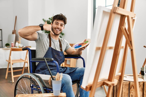 Νεαρός Ισπανόφωνος κάθεται σε αναπηρική καρέκλα ζωγραφική στο στούντιο τέχνης χαμογελώντας κάνει τηλεφωνική χειρονομία με τα χέρια και τα δάχτυλα σαν να μιλάμε στο τηλέφωνο. επικοινωνιακές έννοιες.  - Φωτογραφία, εικόνα