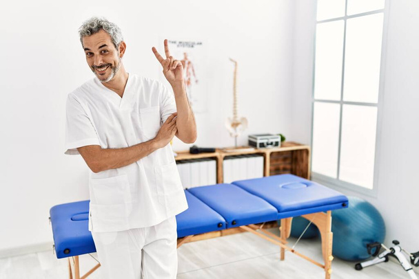 Hombre terapeuta hispano de mediana edad que trabaja en la clínica de recuperación del dolor sonriendo mirando a la cámara mostrando los dedos haciendo señal de victoria. número dos.  - Foto, imagen