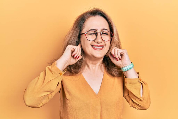 Mujer caucásica de mediana edad que usa ropa casual y gafas que cubren las orejas con los dedos con expresión molesta por el ruido de la música alta. concepto de sordo.  - Foto, imagen