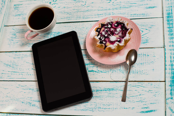 A digital tablet, a cup of coffee and a cake basket with berries and cottage cheese on a white and blue vintage table. Morning breakfast background. Flatley coffee break layout. - Foto, imagen