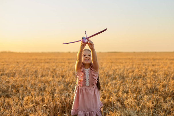 Счастливый ребенок, девочка играет с игрушечным самолетом на летнем пшеничном поле. Маленькая дочка мечтает летать. Беззаботный ребенок играет на открытом воздухе - Фото, изображение
