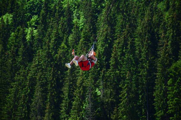A young man in a helmet jumped from bungee jumping and now hangs on a rope, swinging and filming himself on a sports video camera against a blurred background of a green forest - Photo, Image