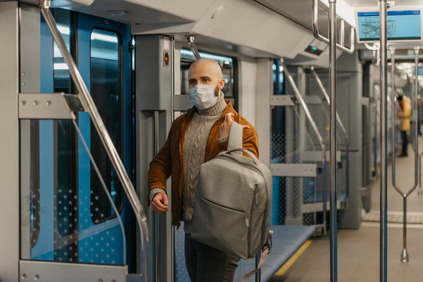Чоловік з бородою в масці для обличчя, щоб уникнути поширення коронавірусу, одягає рюкзак під час їзди на машині метро. Лисий хлопець у хірургічній масці тримає соціальну дистанцію на потязі
. - Фото, зображення