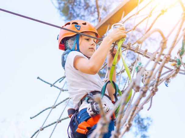 Kid in orange helmet climbing in trees on forest adventure park. Girl walk on rope cables and high suspension bridge in adventure summer city park. Extreme sport equipment helmet and carabiner - Fotografie, Obrázek