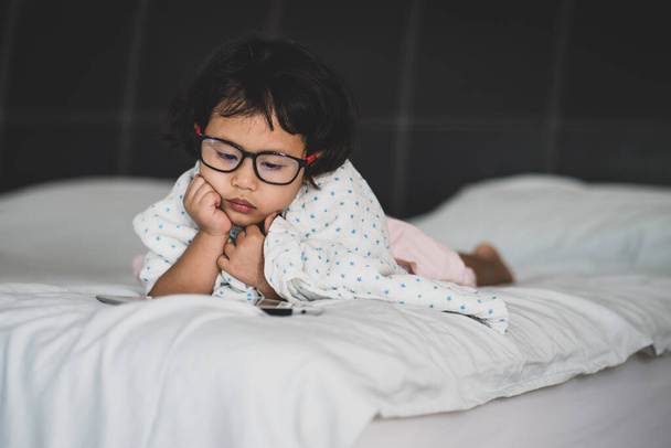 A cute Asian female child with glasses reading a book on a bed - Fotó, kép