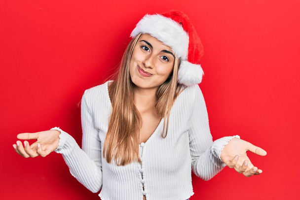 Όμορφη Ισπανίδα γυναίκα φορώντας χριστουγεννιάτικο καπέλο ανίδεη και μπερδεμένη έκφραση με χέρια και χέρια σηκωμένα. έννοια της αμφιβολίας.  - Φωτογραφία, εικόνα