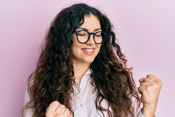 Young brunette woman with curly hair wearing casual clothes and glasses excited for success with arms raised and eyes closed celebrating victory smiling. winner concept.  - Fotoğraf, Görsel