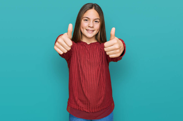 Beautiful brunette little girl wearing casual sweater approving doing positive gesture with hand, thumbs up smiling and happy for success. winner gesture.  - Fotoğraf, Görsel