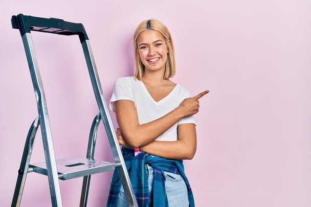 Beautiful blonde woman standing by ladder smiling cheerful pointing with hand and finger up to the side  - 写真・画像