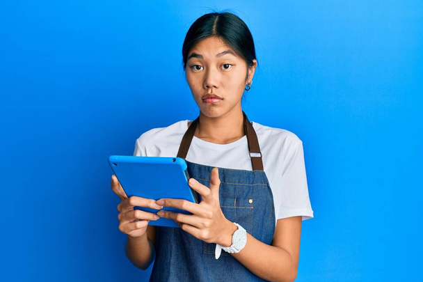 Νεαρή Κινέζα γυναίκα φορώντας ποδιά μάγειρας χρησιμοποιώντας touchpad ανίδεη και μπερδεμένη έκφραση. έννοια της αμφιβολίας.  - Φωτογραφία, εικόνα