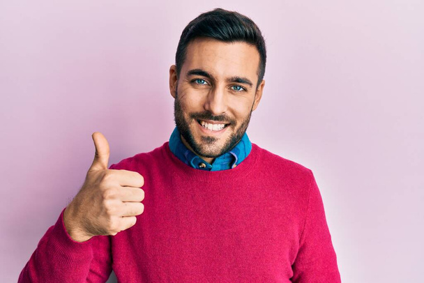 Junger hispanischer Mann in lässiger Kleidung macht glückliche Daumen nach oben Geste mit der Hand. Zustimmender Blick in die Kamera zeigt Erfolg.  - Foto, Bild