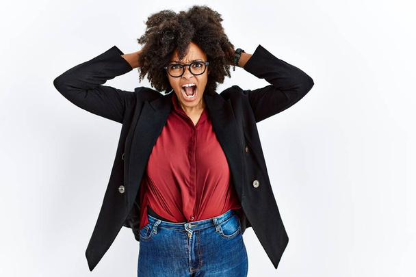 Африканская американка с афроволосами в деловой куртке и очках сумасшедшая и напуганная руками на голове, напуганная и удивленная шоком с открытым ртом  - Фото, изображение