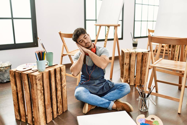 Joven hombre hispano sentado en el estudio de arte durmiendo cansado soñando y posando con las manos juntas mientras sonríe con los ojos cerrados.  - Foto, Imagen