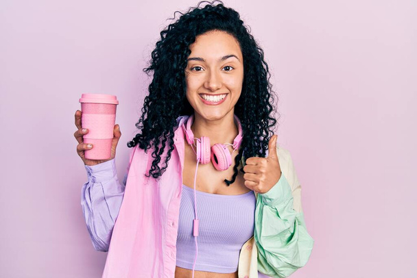 Young hispanic woman with curly hair wearing sportswear and headphones drinking cup of coffee smiling happy and positive, thumb up doing excellent and approval sign  - Photo, Image